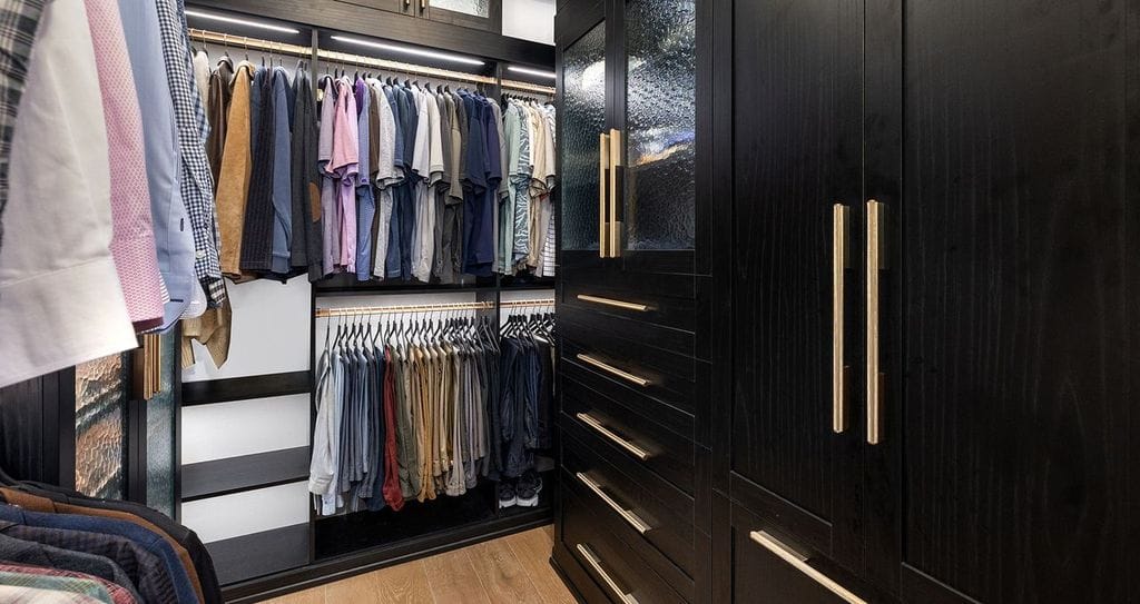 His side of the his and hers closet in a dark wood grain finish with gold hardware, showcasing custom shelving, a built in dresser and led lighting by California Closets
